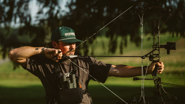 A man in a forest setting drawing a compound bow, wearing a green cap with 'GBB' branding, a dark brown t-shirt, and a black chest rig with binoculars.